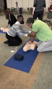 A students places her hands at the elbows of another student who is performing chest compressions. His hands are directly beneath his shoulder and his arms extended as he does the compressions.